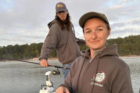  Woman poses for camera with man behind her on the lake.